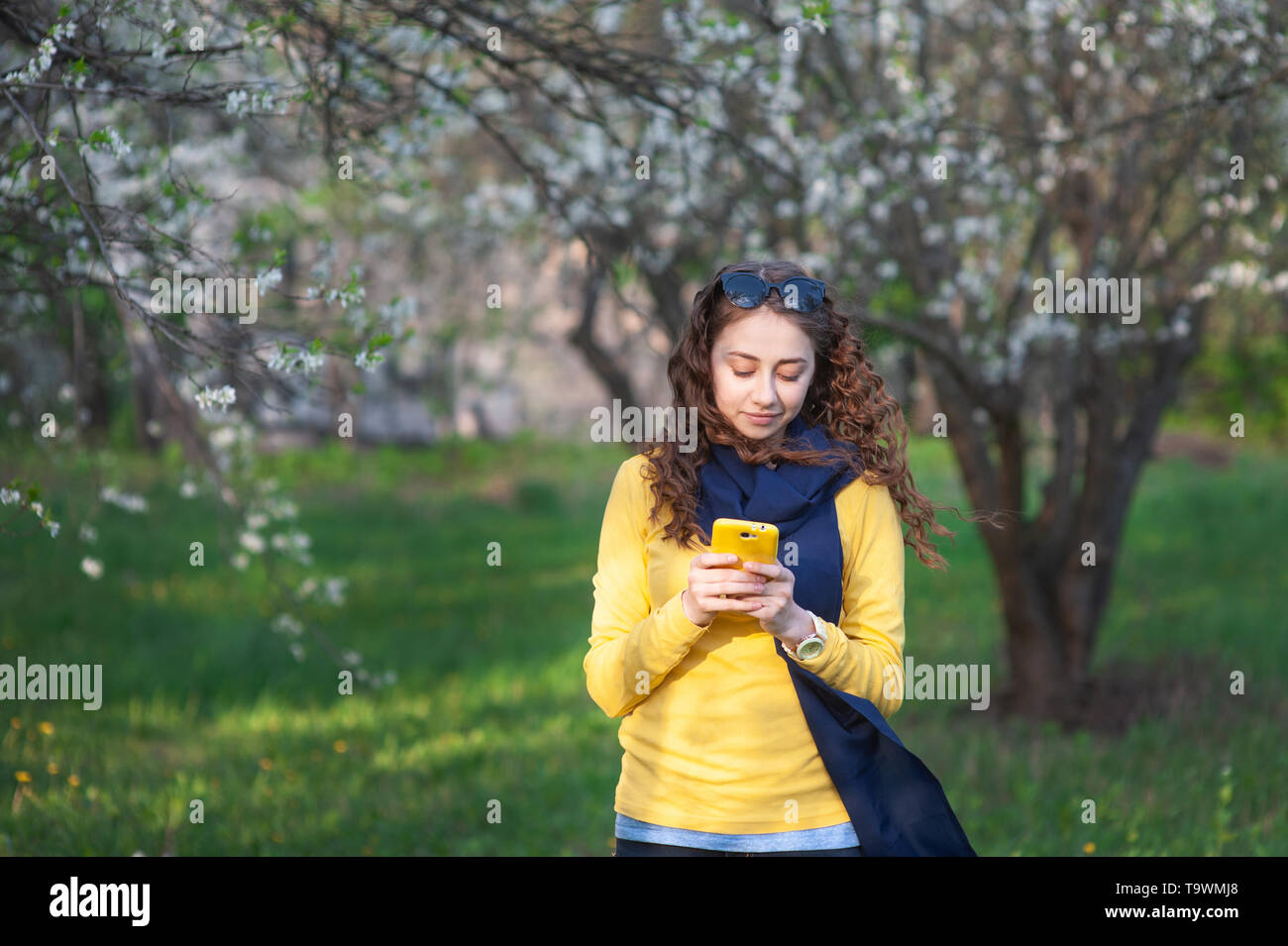 Smilling giovane donna in piedi in un giardino fiorito e scrive sul telefono cellulare. Dei ciliegi. Ritratto di donna bella Foto Stock