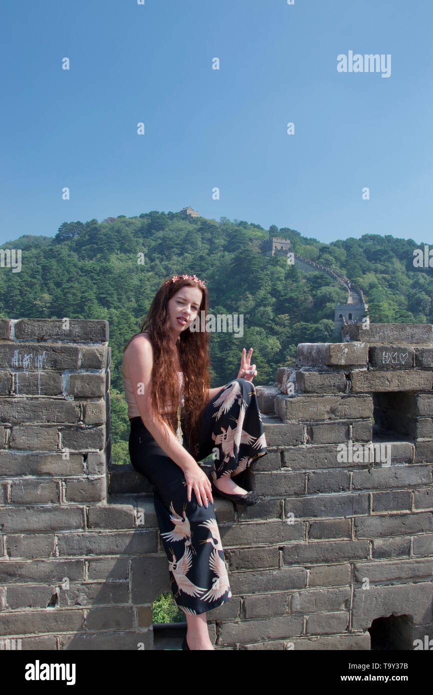 Western caucasian giovane donna indossa turistica di moda modellato pantaloni svasati siede sul parapetto della Grande Muraglia Cinese con la lingua di fuori (soffiaggio Foto Stock