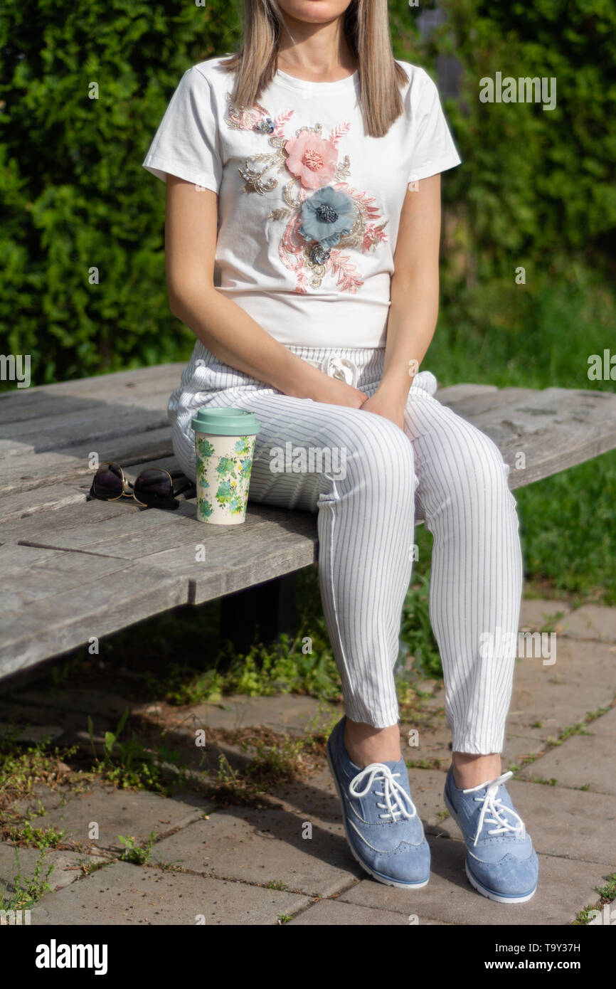 Moda ragazza in pantaloni a righe e scarpe blu seduta su una panca di legno  accanto a una tazza di caffè e occhiali da sole nel parco sulla molla  giornata di sole
