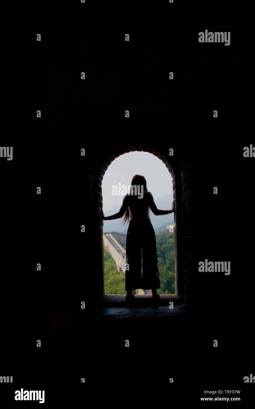 Un capelli lunghi lady indossa pantaloni svasati (Bengala) sorge in un portone, in silhouette, con la Grande Muraglia della Cina dietro di lei. Western turistico in come Foto Stock
