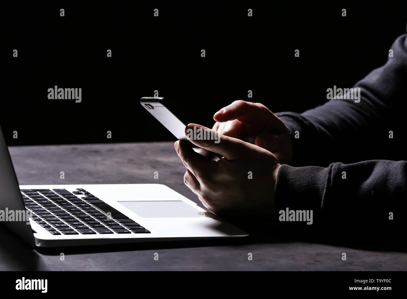 Professional hacker con il computer portatile e il telefono cellulare seduta a tavola su sfondo scuro, primo piano Foto Stock