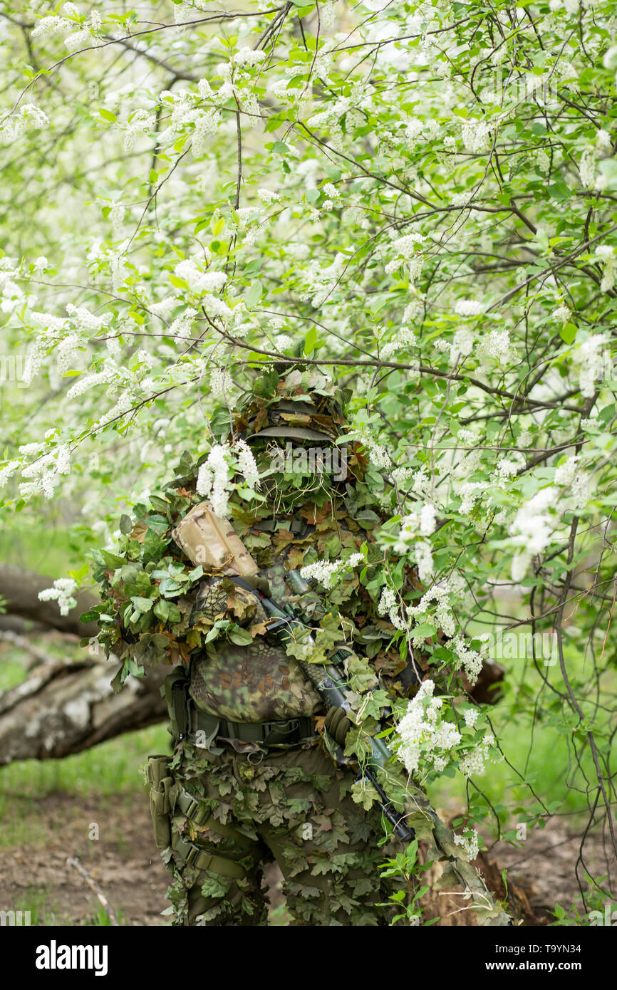 Simpatico ritratto di un maschio militare sniper in camuffamento, maschera, cappello e fucile sulla sua mano contro lo sfondo di una fioritura di ciliegio in estate fuori Foto Stock