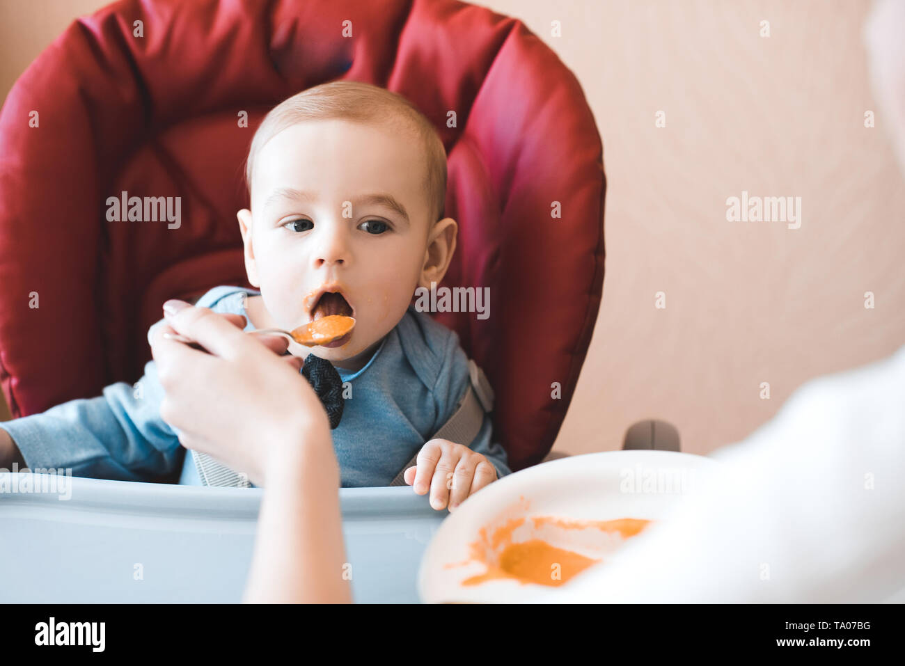 Baby boy 1 anno vecchio mangiare purea di mela seduto nella sedia in cucina. La madre del bambino di alimentazione. Infanzia. Foto Stock