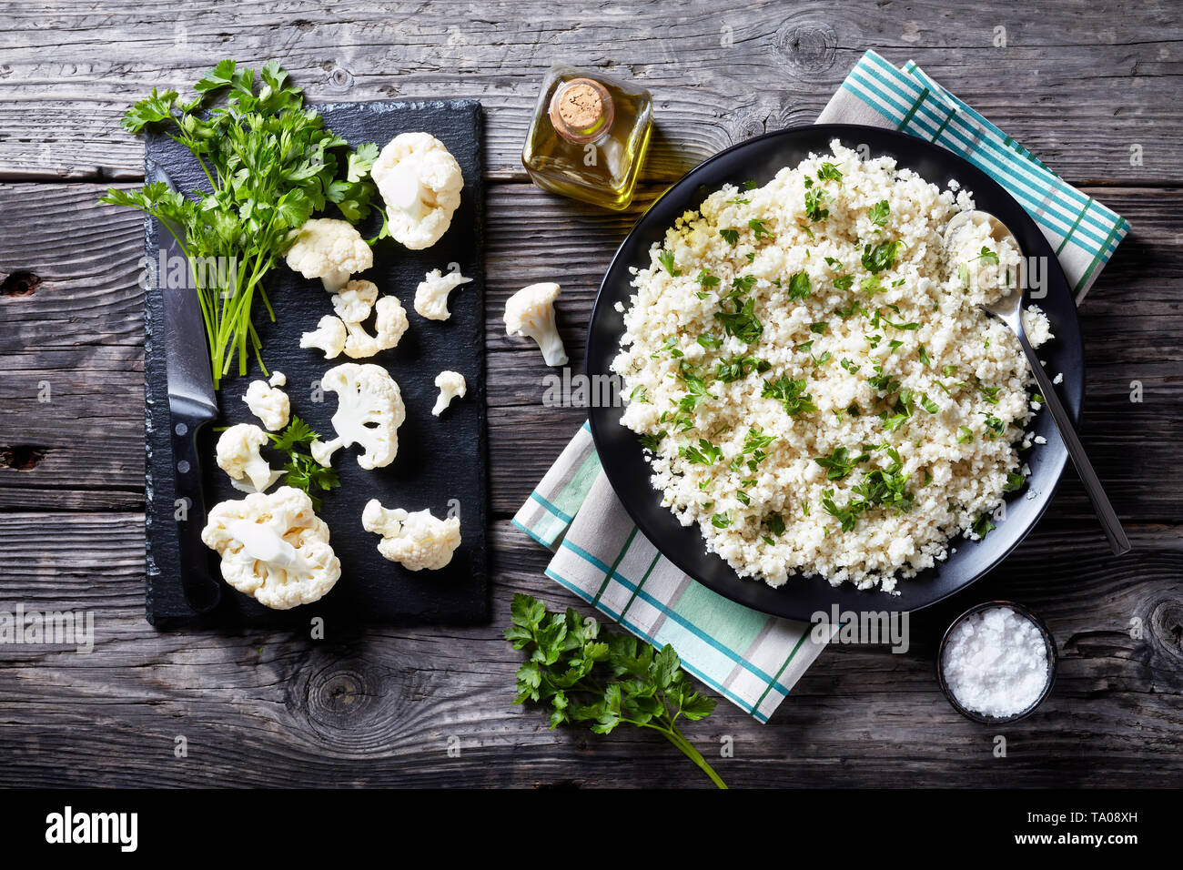 Vista aerea del cavolfiore riso o cous cous mescolato con del prezzemolo tritato finemente in una ciotola nero su un vecchio tavolo rustico con ingredienti su un taglio b Foto Stock