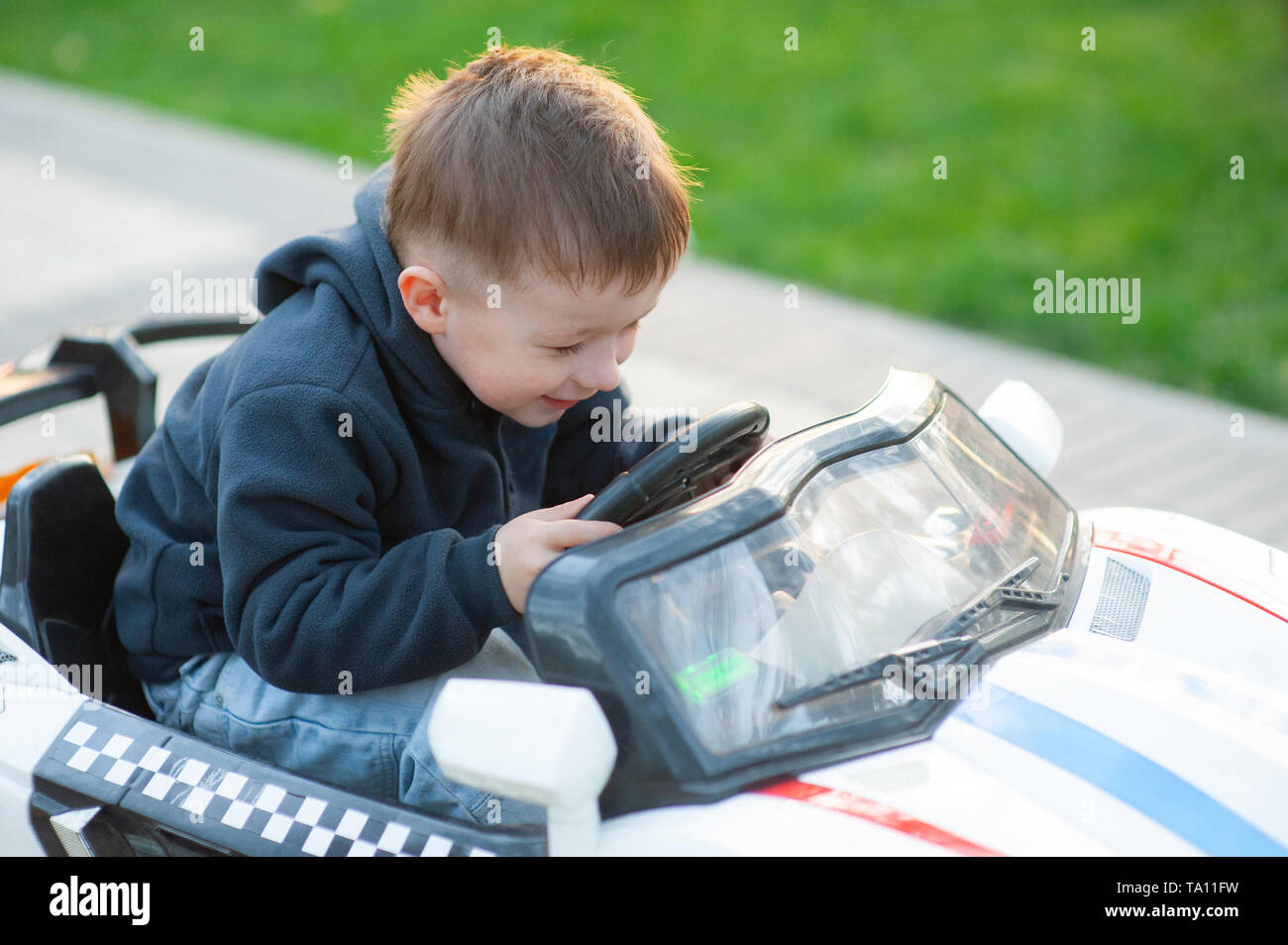 Carino piccolo ragazzo la guida meccanica auto giocattolo girando il volante con uno sguardo di concentrazione come suona in un parco della città Foto Stock