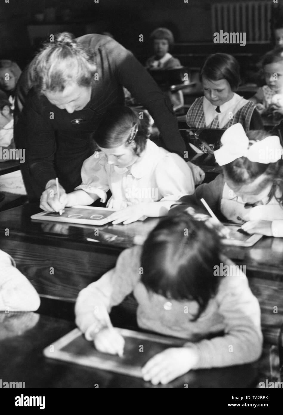 La classe di un GIR'ls classe siedono nei banchi di scuola e di scrivere con il gesso sulle loro schede di scrittura. Un insegnante è aiutare una delle ragazze. Foto Stock