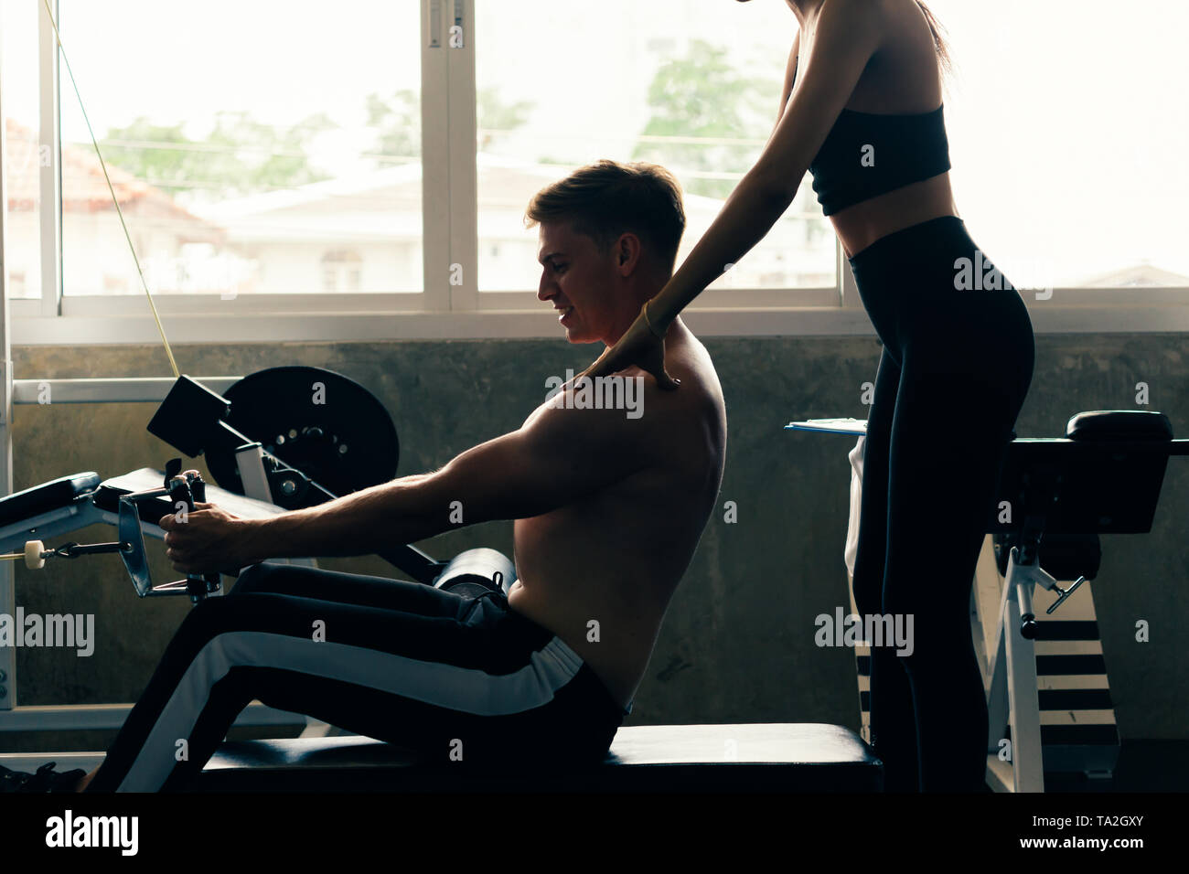 Giovane uomo caucasico in maglia bianca in una palestra, allenamento duro tirando i pesi in seduto cavo macchina di riga con il supporto dalla femmina personal trainer Foto Stock