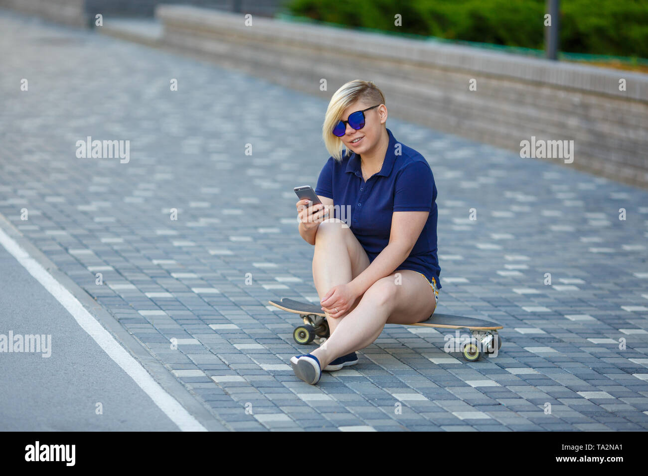 Elegante ragazza di grandi dimensioni, taglio di capelli alla moda in vetri a specchio, seduto su un longboard contro lo sfondo dell'edificio, tenendo il telefono cellulare Foto Stock