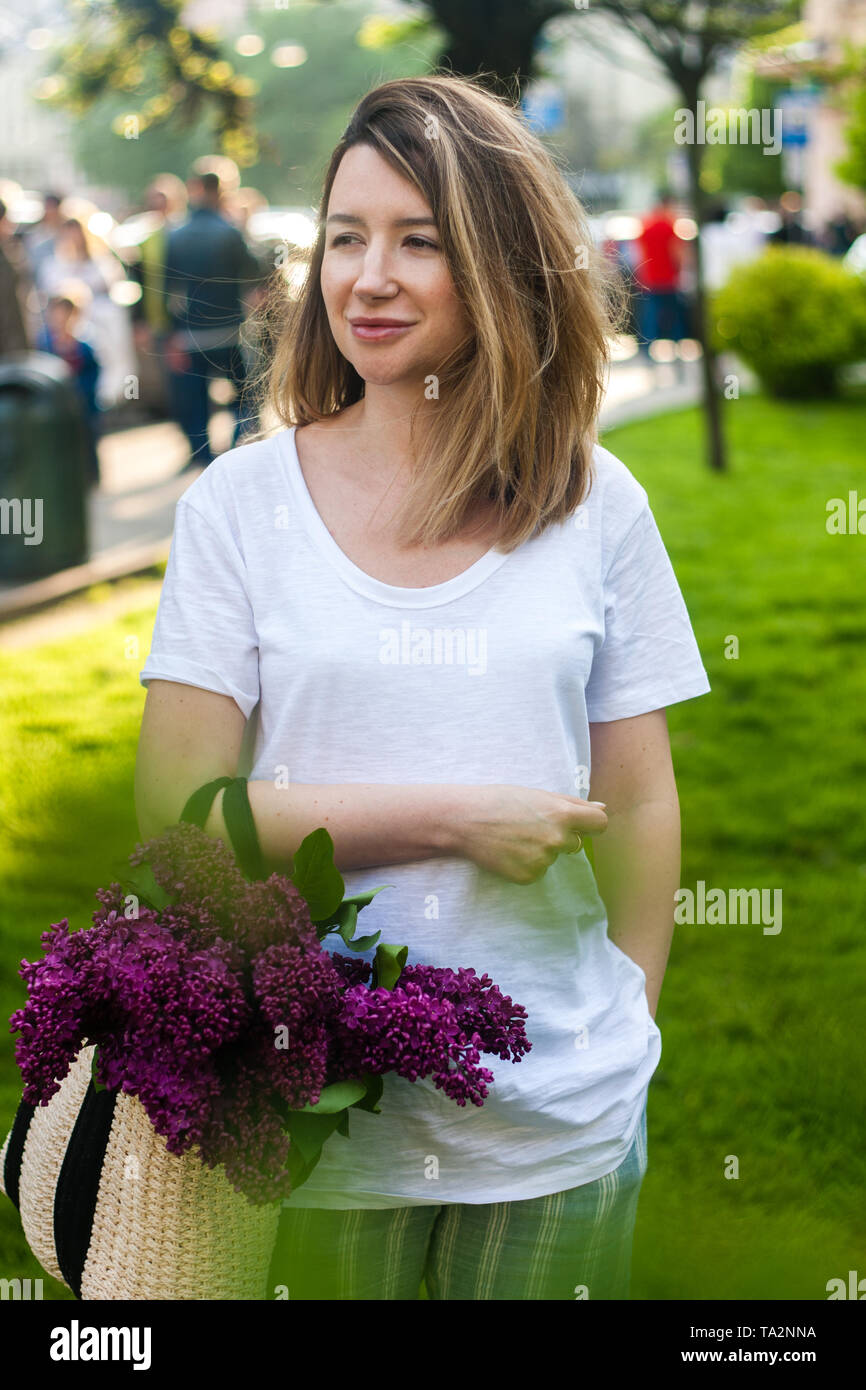 Donna elegante azienda borsa di paglia con un vivido mazzetto di fiori lilla. Verde brillante sullo sfondo di erba. Foto Stock