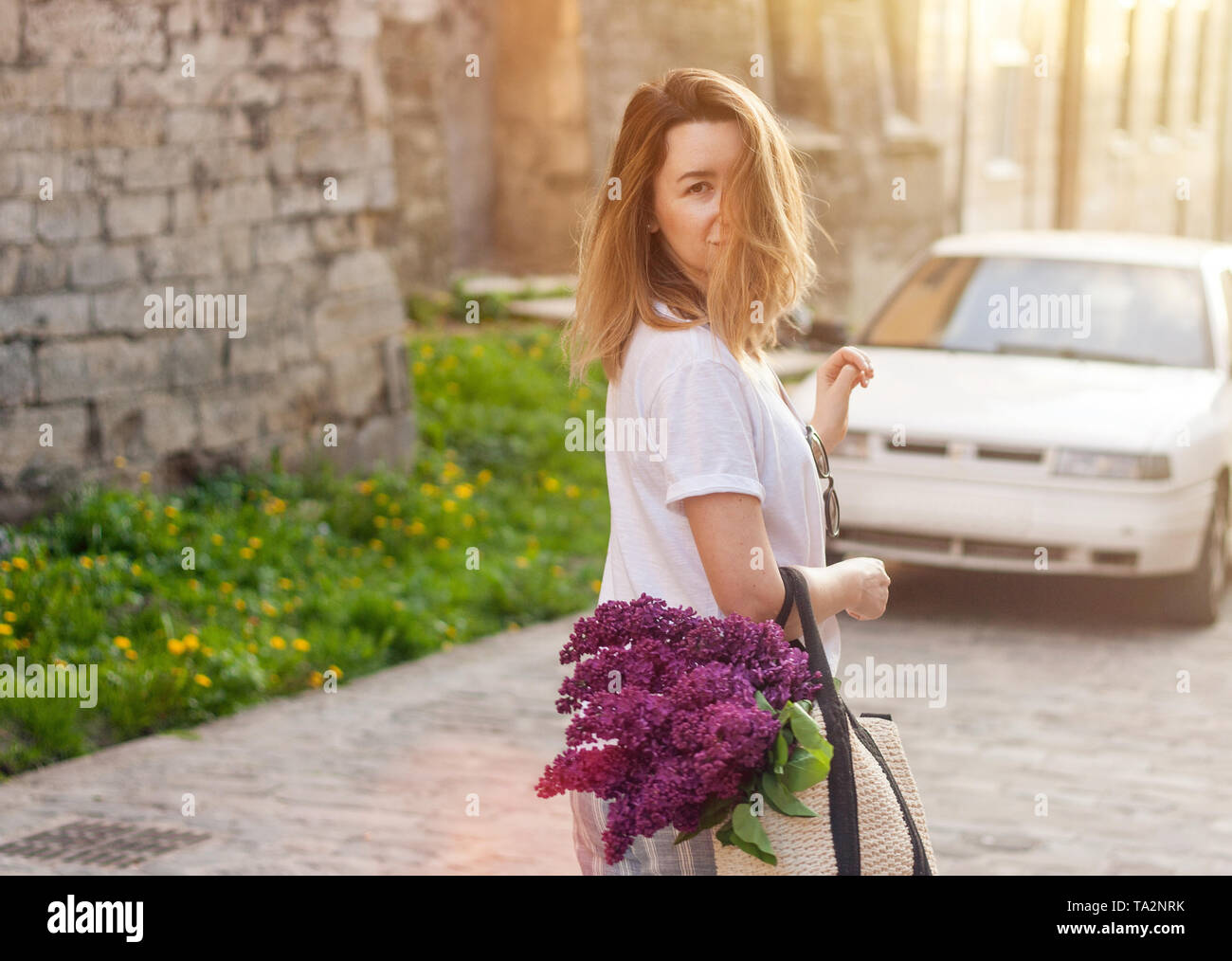 Donna elegante azienda borsa di paglia con un vivido mazzetto di fiori lilla e camminare per la strada. Moody ritratto solare. Foto Stock