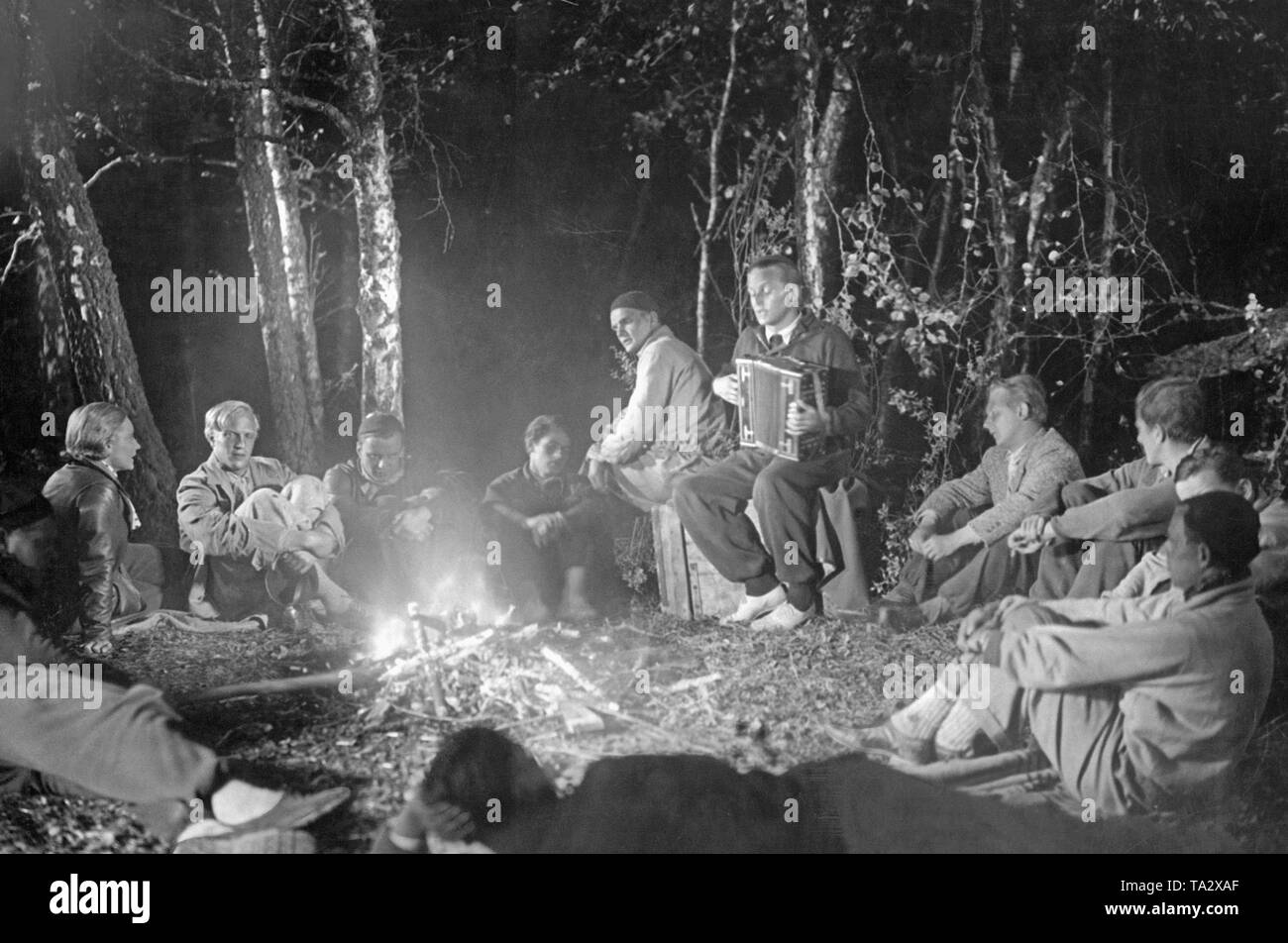 Un "Viaggi in gruppo" siede comodamente in corrispondenza di un fuoco di campo nella foresta, uno degli uomini più giovani sta suonando la fisarmonica. Foto Stock