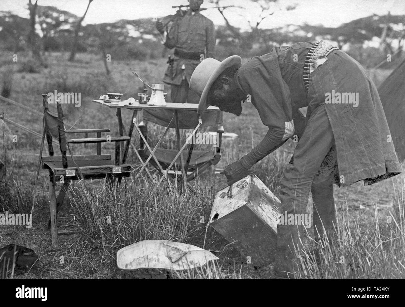Turismo africani in Africa nera: in Africa nera, che è ancora dominato dalle potenze coloniali, un uomo nativo si versa acqua in un recipiente di lavaggio per il big game cacciatori nel mezzo della savana, dietro di lui è un tavolo coperto e in background una pistola portante. Foto Stock