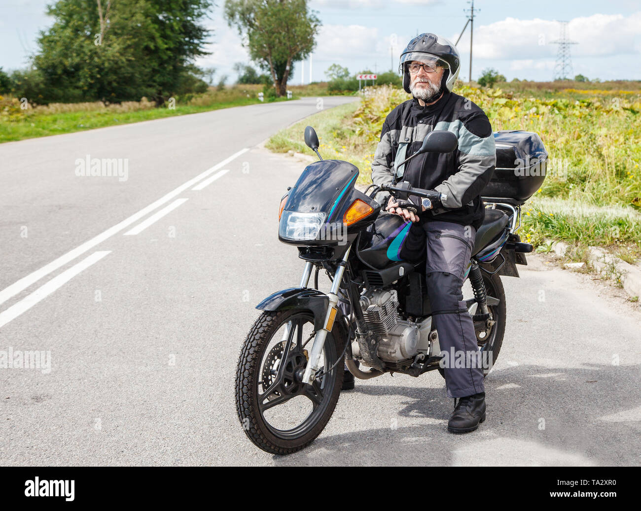 Anziani motociclista che indossa una giacca e occhiali con un casco seduto su una motocicletta sulla strada aperta su soleggiate giornate estive Foto Stock