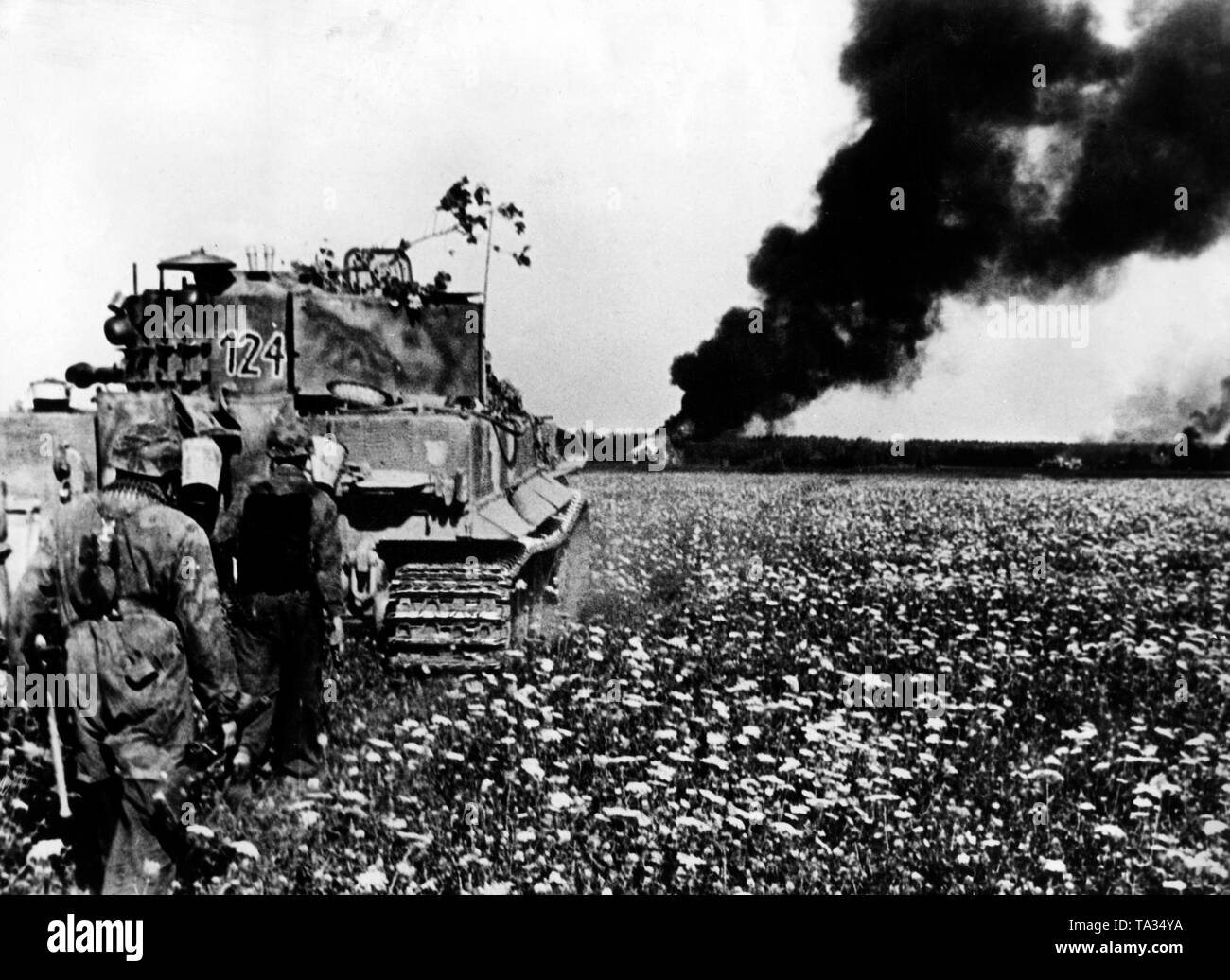 Un tedesco Kampfpanzer VI (Tiger) in un prato in Lituania. Dietro, diversi fanti sono in esecuzione sotto il suo coperchio. Sullo sfondo il fumo di una casa in fiamme. Foto di Propaganda Company (PK): corrispondente di guerra Slickers. Foto Stock