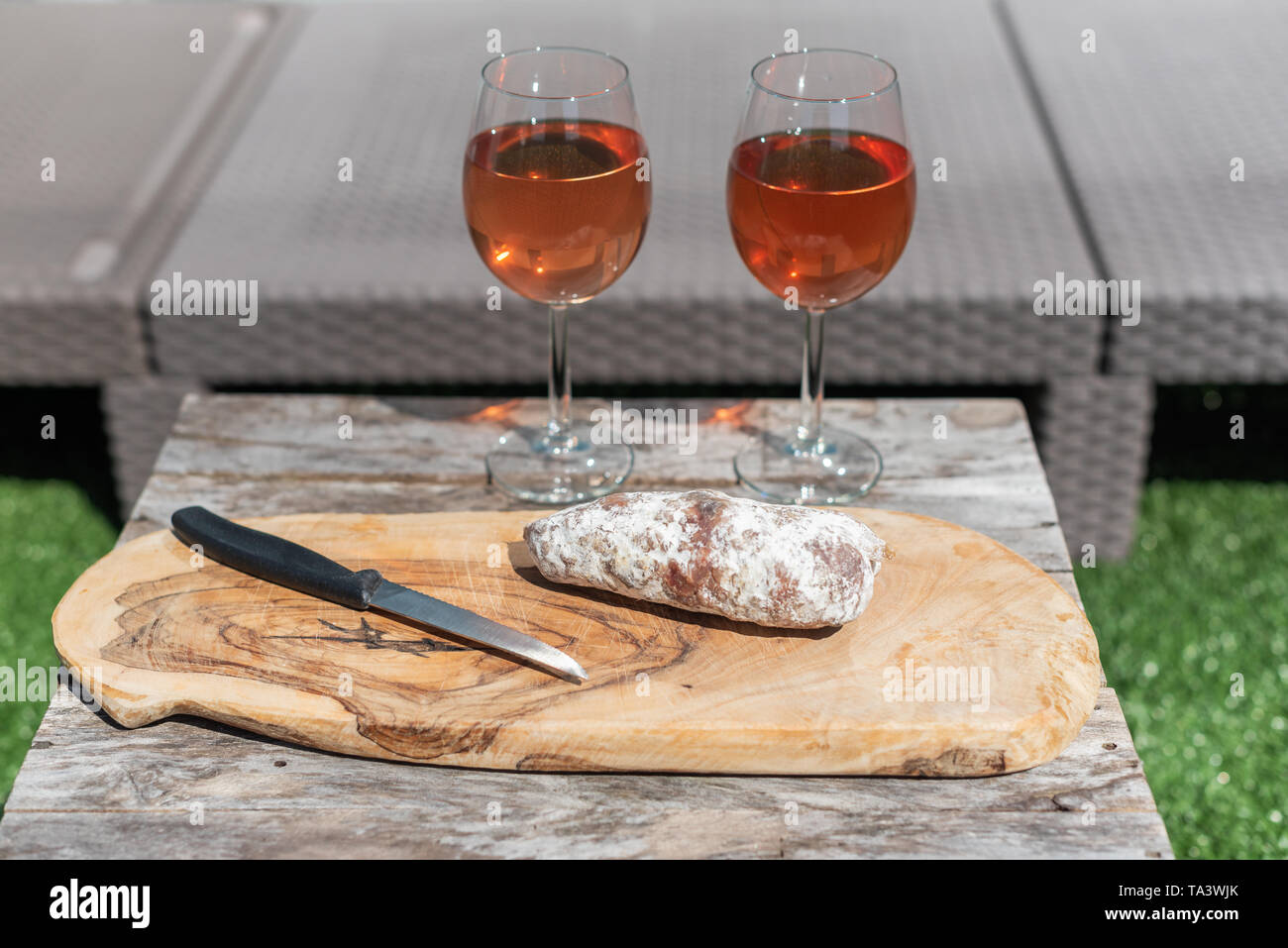 Due bicchieri di vino rosato e una salsiccia secca con un coltello su un tavolo di legno in un giardino in una giornata di sole Foto Stock