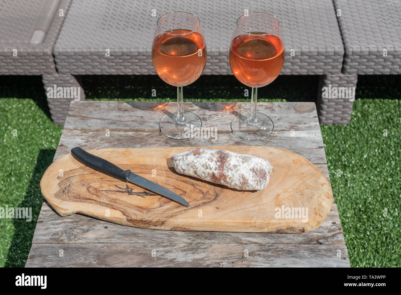 Due bicchieri di vino rosato e una salsiccia secca con un coltello su un tavolo di legno in un giardino in una giornata di sole Foto Stock