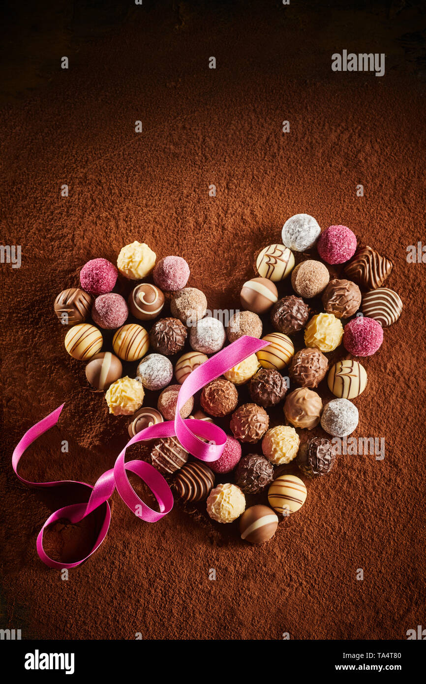 Selezione gourmet di fatto a mano cioccolatini disposti come un cuore su uno sfondo di cacao in polvere con twirled pink ribbon e vignette simbolica dell amore Foto Stock