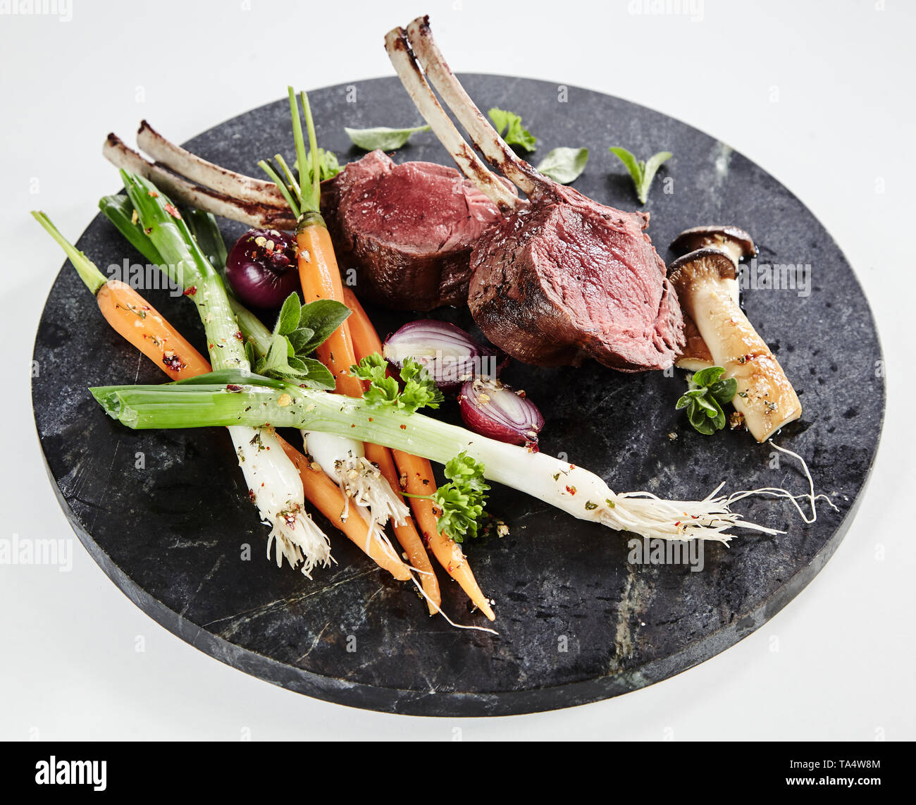 Mezzo raro costolette di agnello con tutta la molla bambino verdure e funghi conditi con erbe e spezie, servito su un round piatto di ardesia Foto Stock