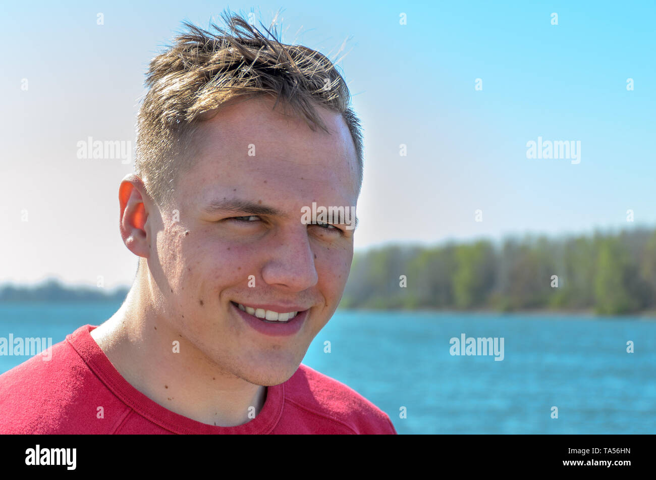 Mezzo giro a busto ritratto di un giovane ragazzo in t-shirt rossa, in piedi all'aperto sul lago o fiume. Guardando la fotocamera e sorridente Foto Stock