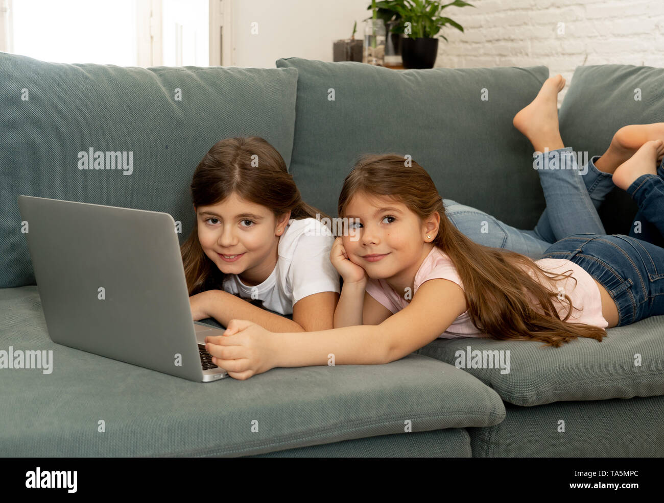 Carino bambina e sorella giocare insieme sorridente e avente il tempo di incollaggio utilizzando un computer portatile sul divano di casa. La famiglia felice fratelli rapporto Foto Stock