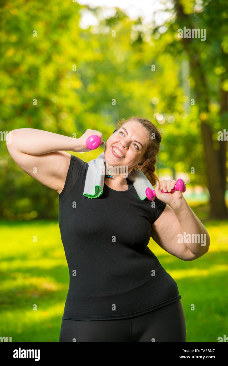 Ritratto verticale di un felice plus-size modello con manubri durante un allenamento in un parco estivo Foto Stock