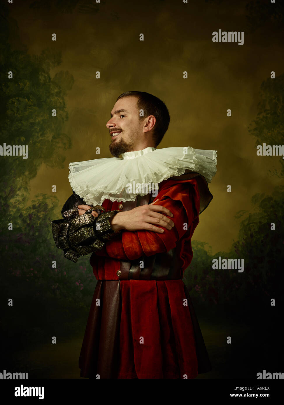 Giovane uomo come un cavaliere medievale su studio scuro dello sfondo. Ritratto in chiave di basso del modello maschile in costume retrò. In piedi e sorridente. Le emozioni umane, co Foto Stock