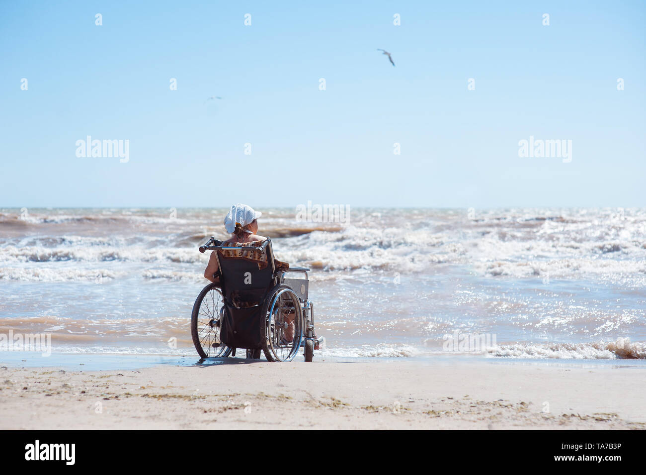 Vista posteriore di una donna disabile seduta su una sedia a rotelle sulla spiaggia in una giornata di sole. Guarda l'oceano Foto Stock