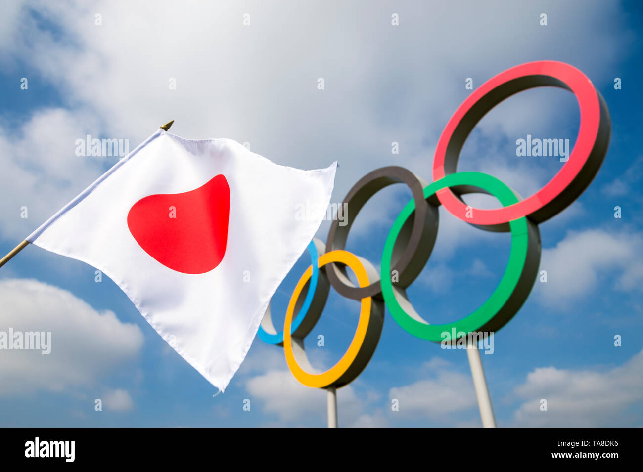 Londra - Aprile 2019: una bandiera giapponese sventola al vento nella parte anteriore di anelli olimpici in piedi sotto il luminoso cielo blu. Foto Stock