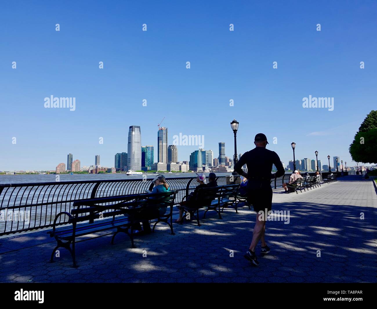 Pareggiatore passando da persone rilassante sulle panchine lungo il fiume Hudson, Batteria Esplanade, New York, NY, STATI UNITI D'AMERICA. Foto Stock