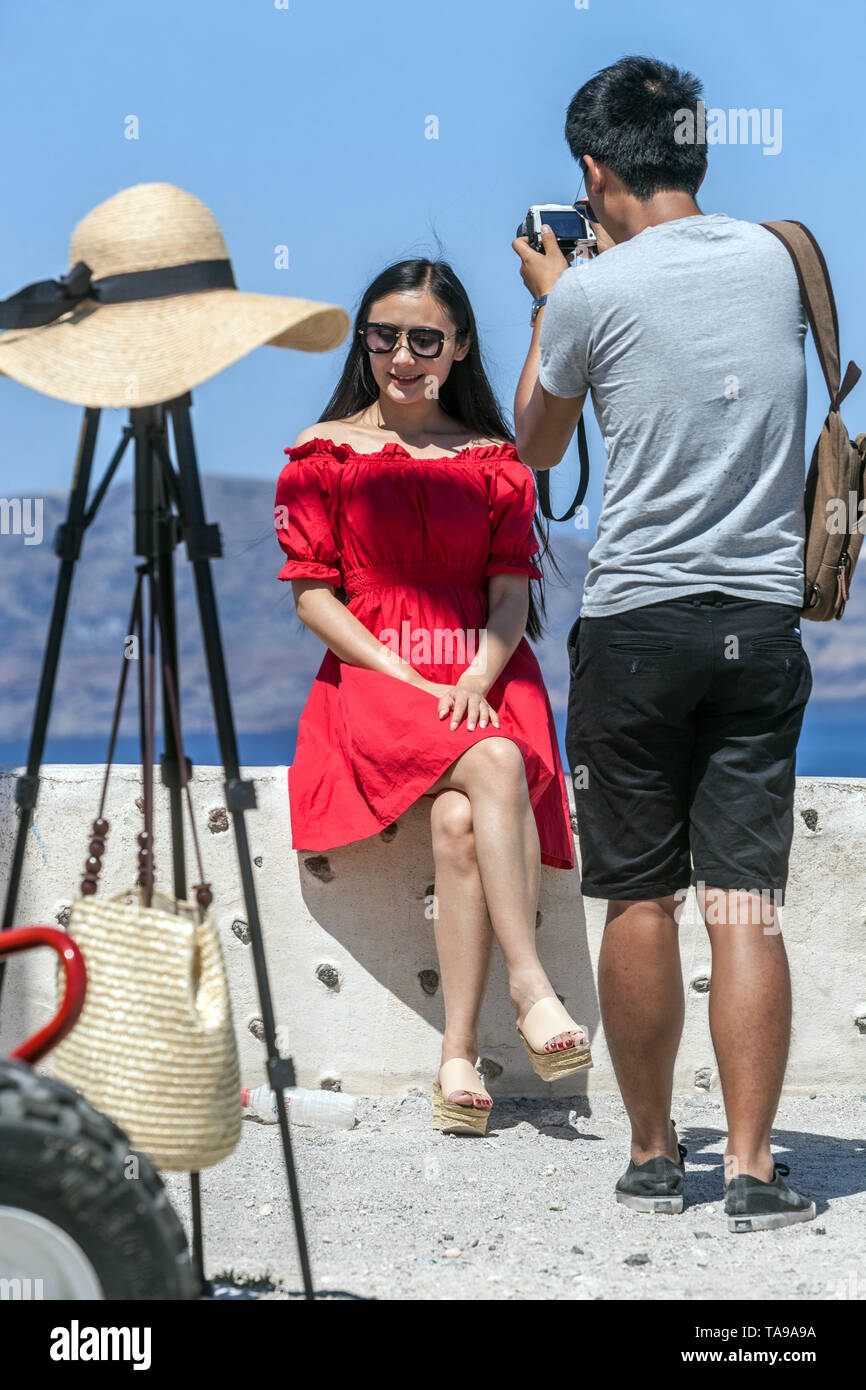 Santorini, turisti asiatici, uomo facendo foto donna in abito rosso al di sopra di mare sulla terrazza, Isole Greche grecia Europa Foto Stock