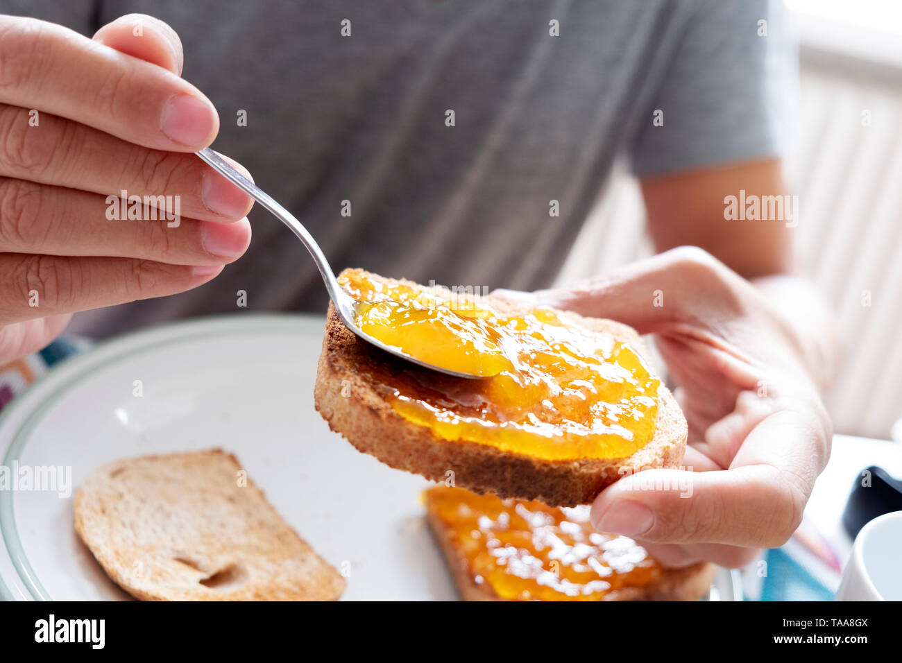 Primo piano di un giovane uomo caucasico, indossando un grigio casual T-shirt, seduti a un tavolo set, diffondendo alcuni arancione o confettura di pesche su un toast Foto Stock