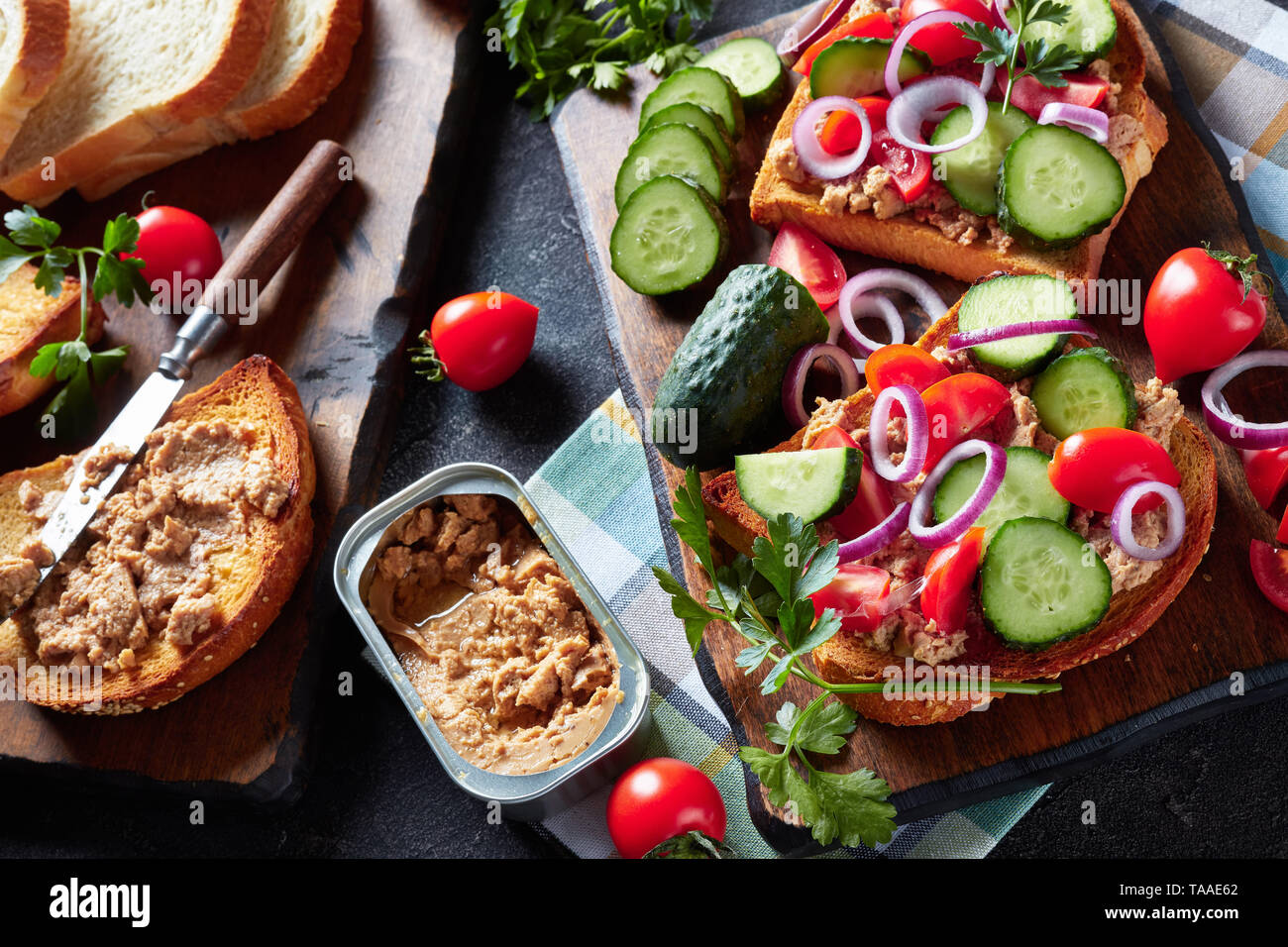 Toast per la stagione estiva la colazione con fegato di merluzzo pate, guarnita con fette di cetriolo, pomodoro e cipolla rossa anelli su di un tagliere con ingredienti su un ki Foto Stock