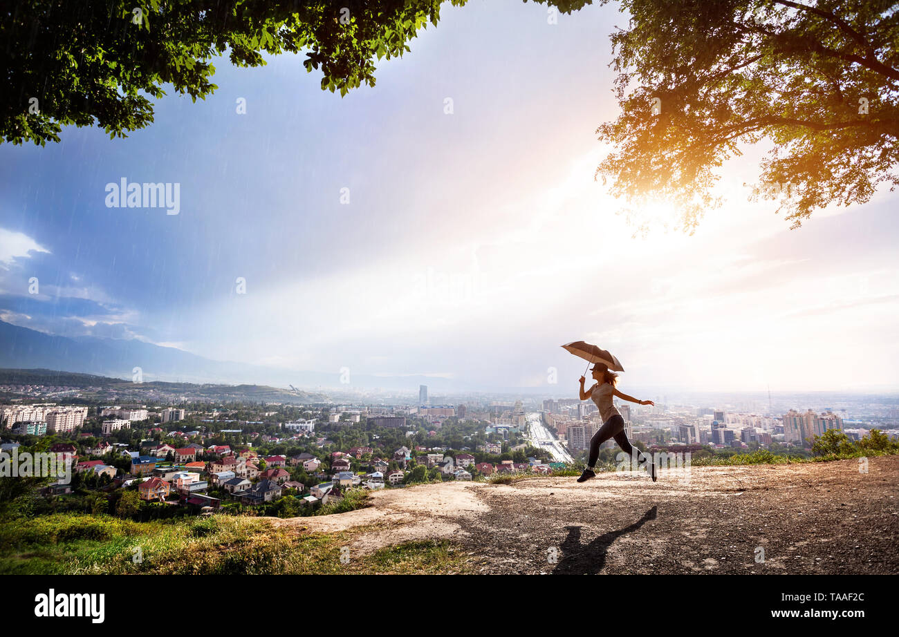 Silhouette di donna con ombrello saltando la vista del panorama cittadino a rainy tramonto spettacolare. Foto Stock