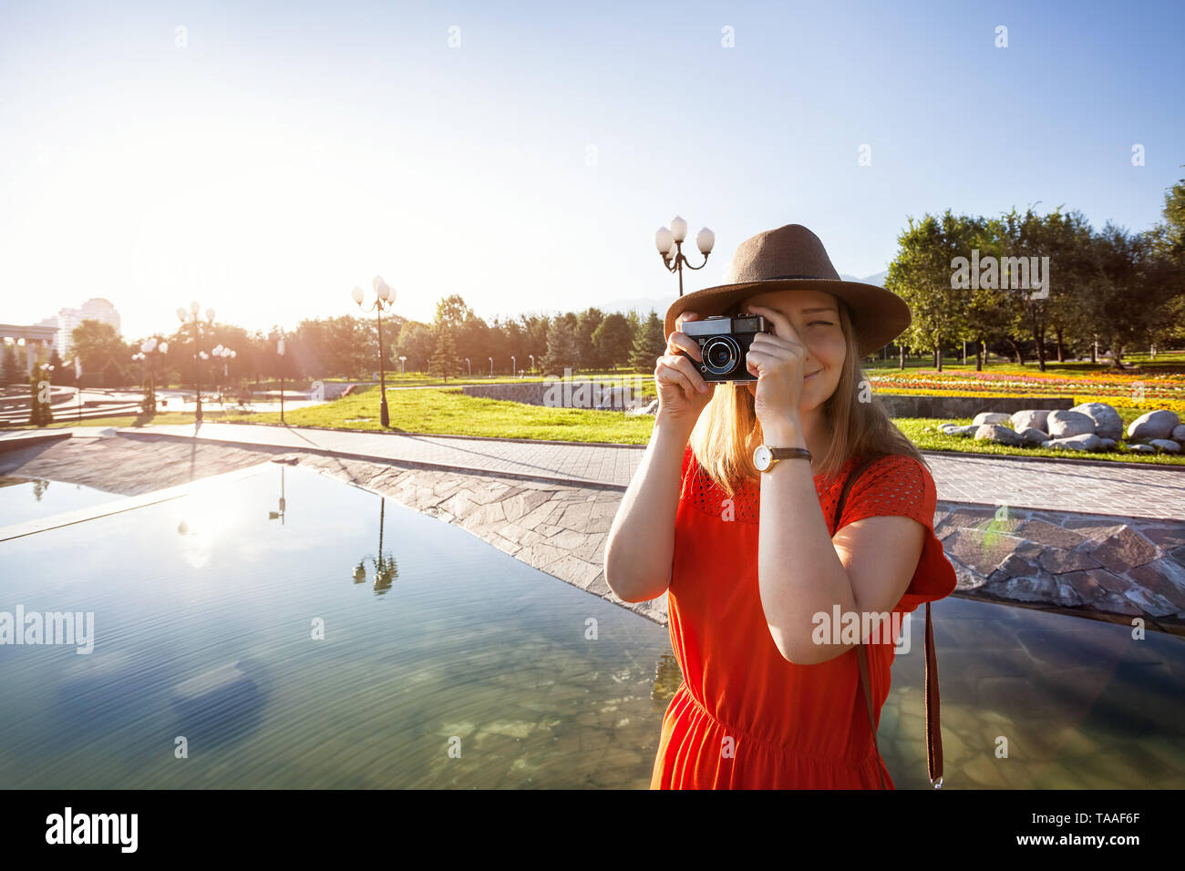 Felice donna turistica nel cappello e abito arancione con fotocamera vintage nel parco di sunrise Foto Stock