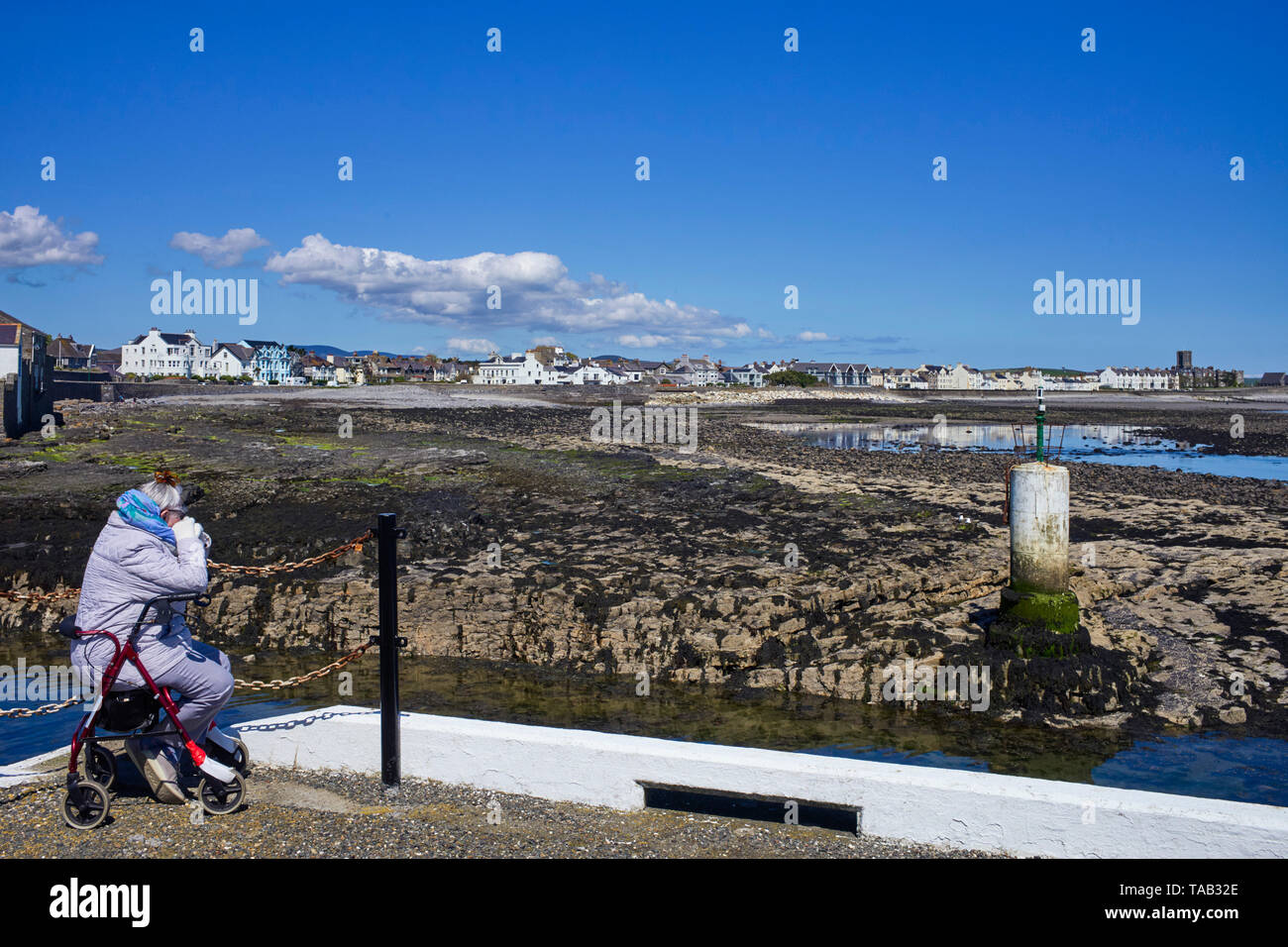 Donna anziana indossando giacca imbottita seduta dal lato del porto esterno muro in Castletown, Isola di Man affacciato sulla baia con la bassa marea Foto Stock