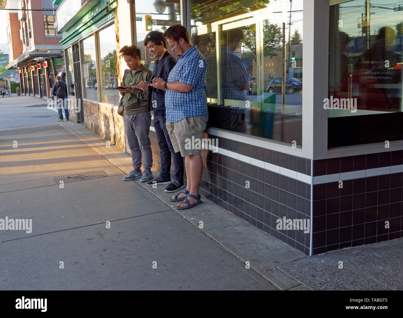 Tre giovani uomini usando i loro cellulari mentre si sta in piedi sul marciapiede, Main Street, Vancouver, BC, Canada Foto Stock