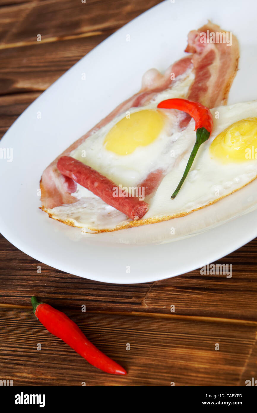 Uova fritte con pancetta, peperoncino rosso e piccole salsicce giacciono su una piastra bianca su un naturale piano di legno di tavole di pino. La luce diurna. L'immagine verticale. Foto Stock
