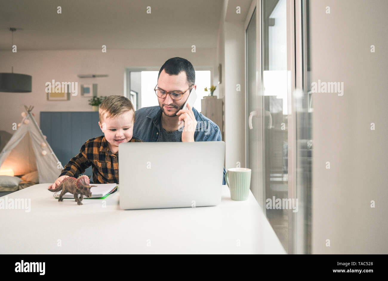Padre lavora a tavola in casa ufficio con Figlio seduto sul suo giro Foto Stock