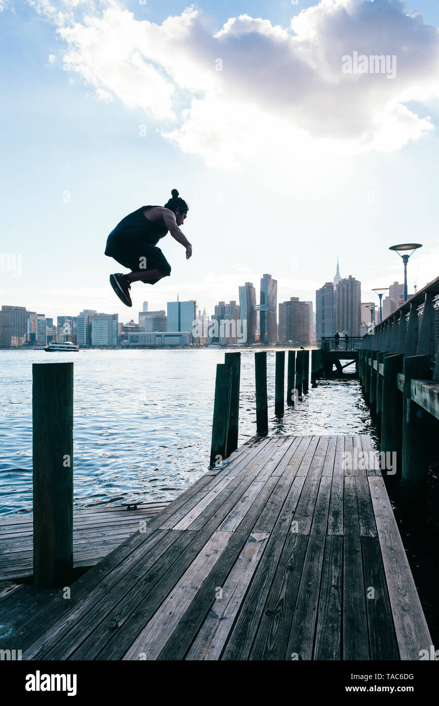 Stati Uniti d'America, New York, Brooklyn, il giovane uomo che fa parkour saltare da un palo di legno nella parte anteriore dello skyline di Manhattan Foto Stock