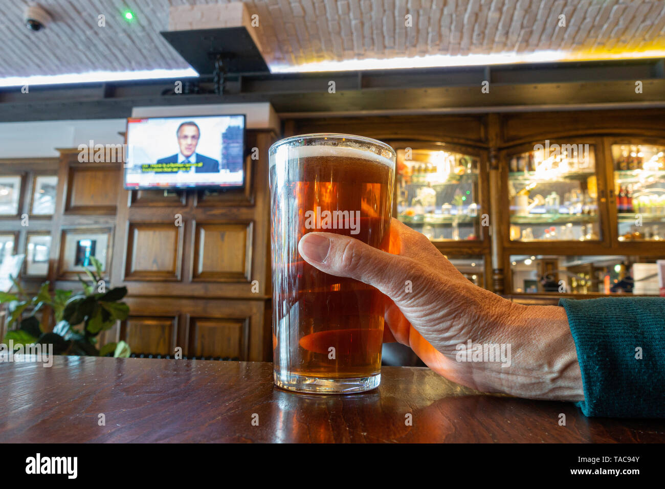 Persona in possesso di una pinta di birra artigianale in pub con notizie sulla TV in background. Regno Unito Foto Stock