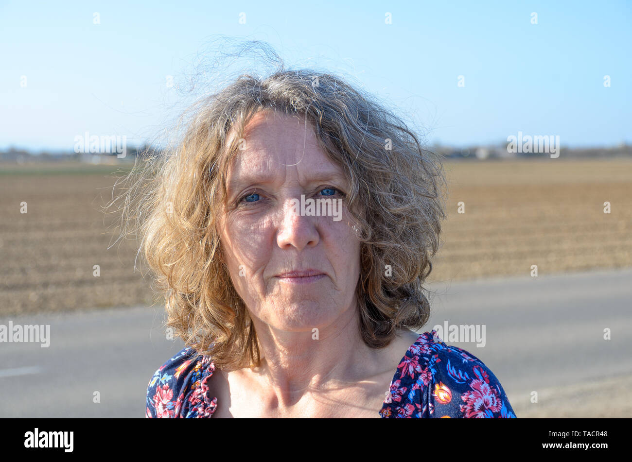 Premurosa donna di mezza età con spallamento Ricci Lunghezza capelli in piedi all'aperto in campagna fissando attentamente la fotocamera A Foto Stock Premurosa donna di mezza età con spallamento Ricci Lunghezza capelli in piedi all'aperto in campagna fissando attentamente la fotocamera A Foto Stock