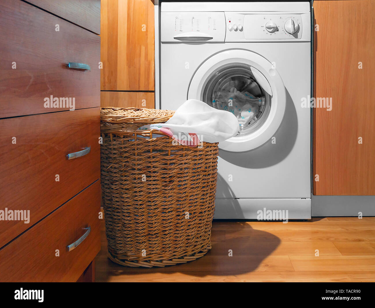 In vimini grande cesto per la biancheria, coperchio aperto, vicino alla  macchina lavabiancheria a carica frontale con lavanderia. Interno di una  casa locale lavanderia. Interni in legno di Design Foto stock 