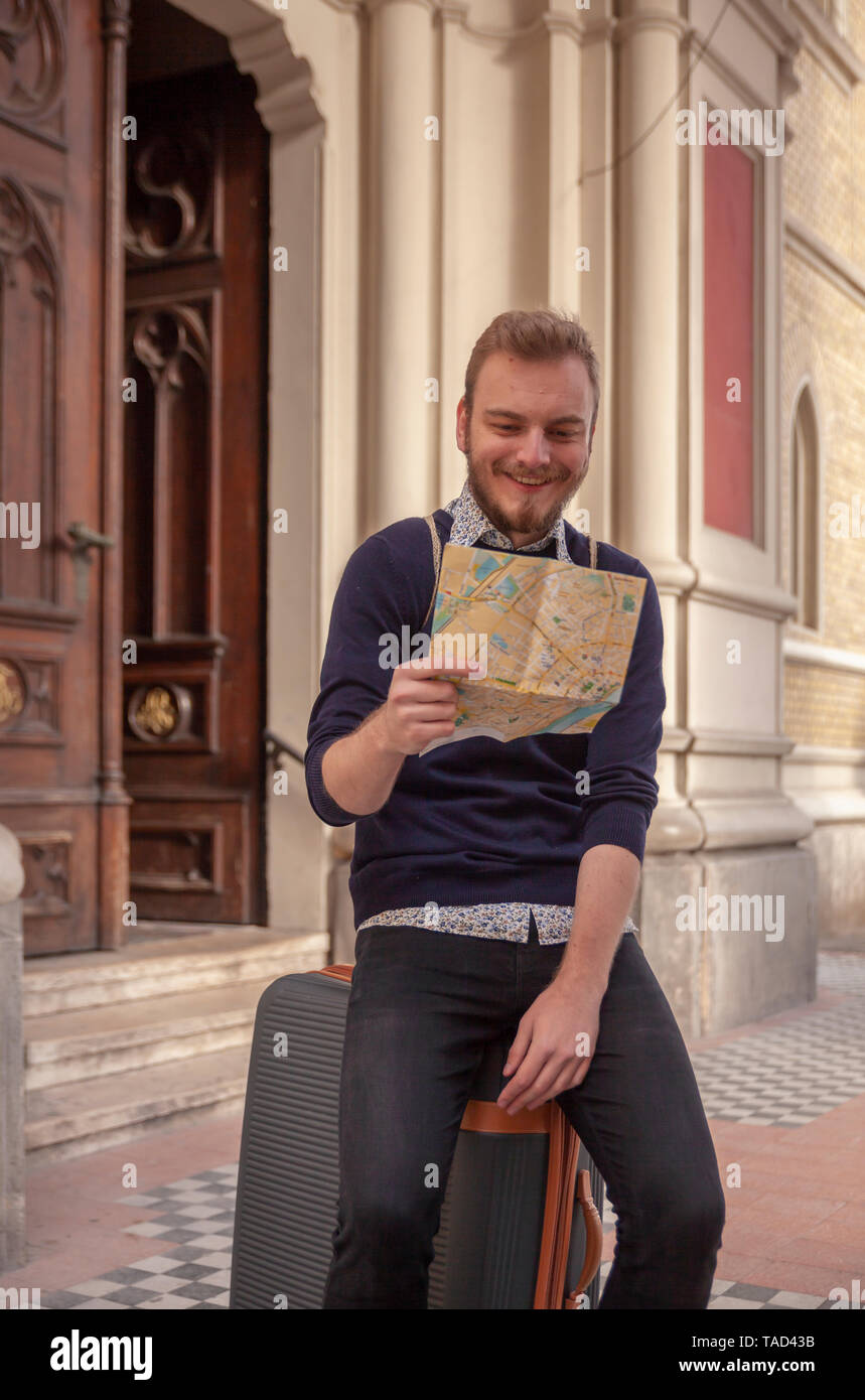 Un giovane uomo - traveler o turistico, guardando la mappa della città mentre è seduto su una valigia. rilassante in una vecchia architettura europea stile, all'esterno. Foto Stock