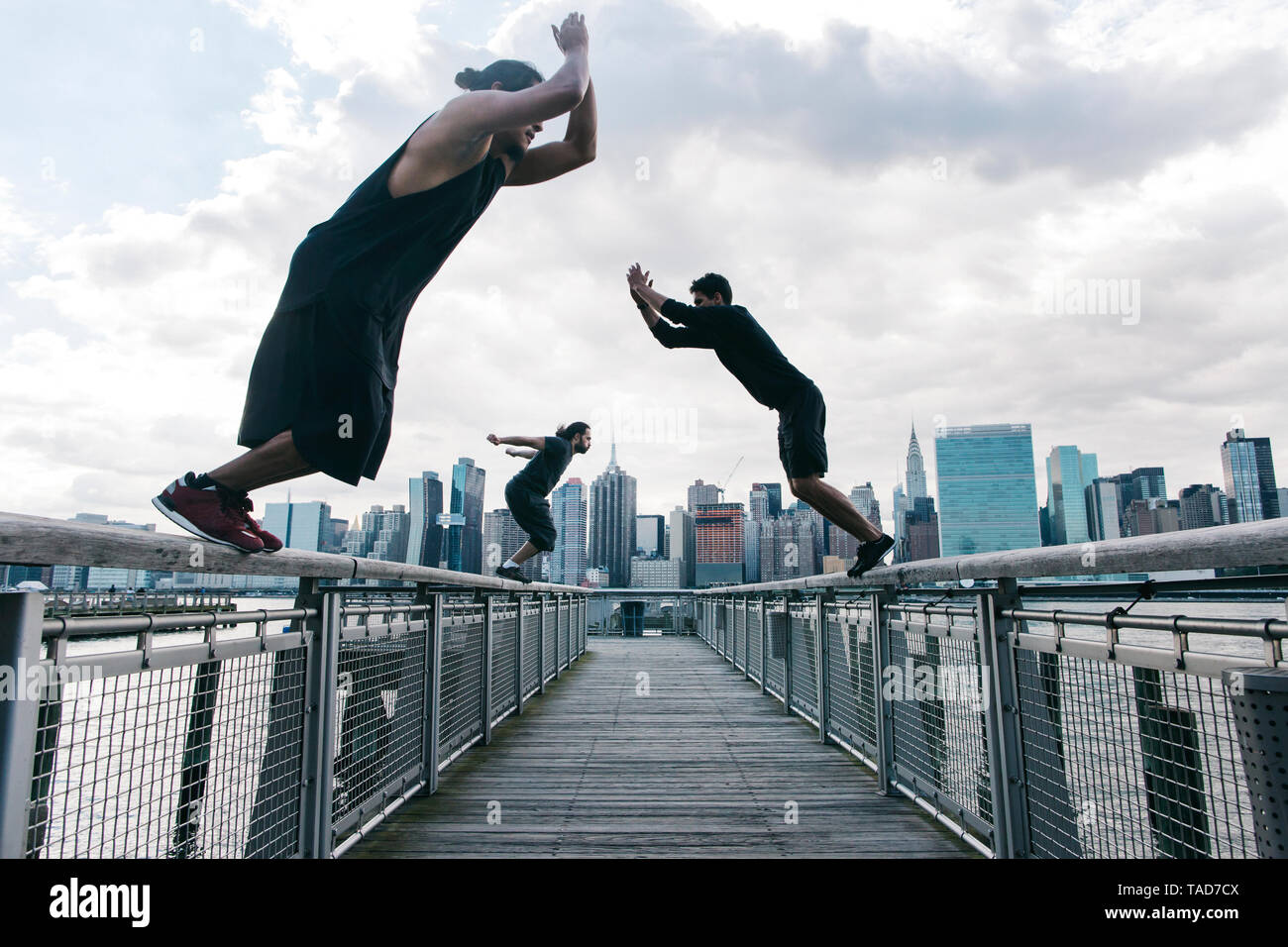 Stati Uniti d'America, New York, Brooklyn, tre giovani uomini a fare parkour salta sul molo di fronte skyline di Manhattan Foto Stock