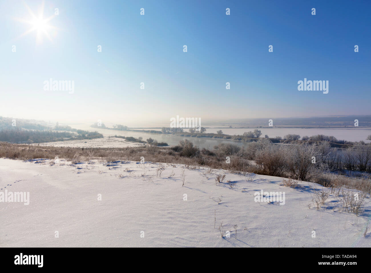Rurali paesaggio invernale, i raggi del sole in un grado di velatura del cielo blu giacciono su un fiume di avvolgimento e coperti di neve i campi, alberi e cespugli. Foto Stock
