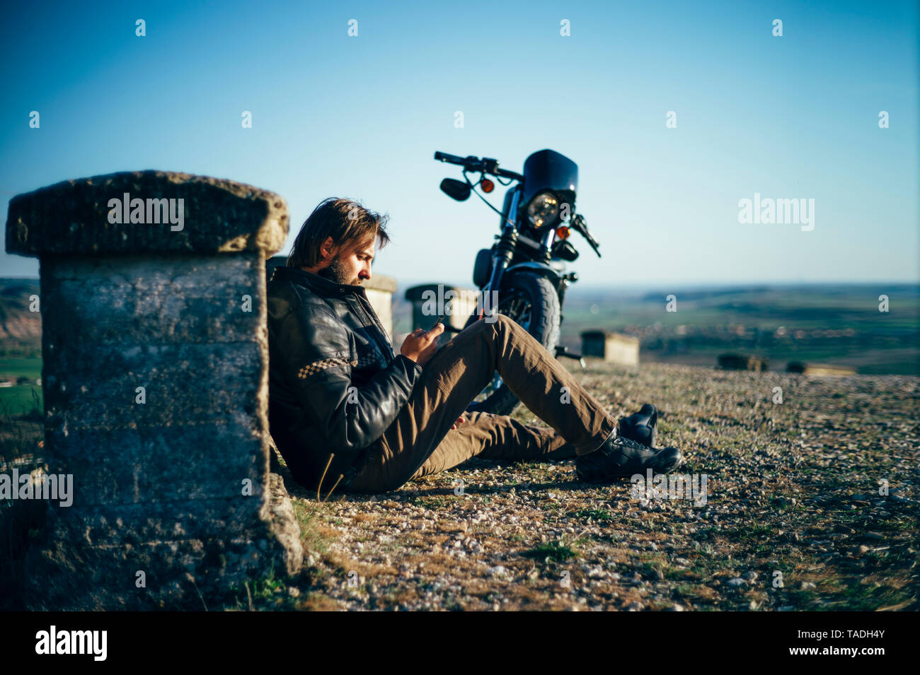 Uomo con custum motociclo avente una pausa seduta sul terreno tramite telefono cellulare Foto Stock