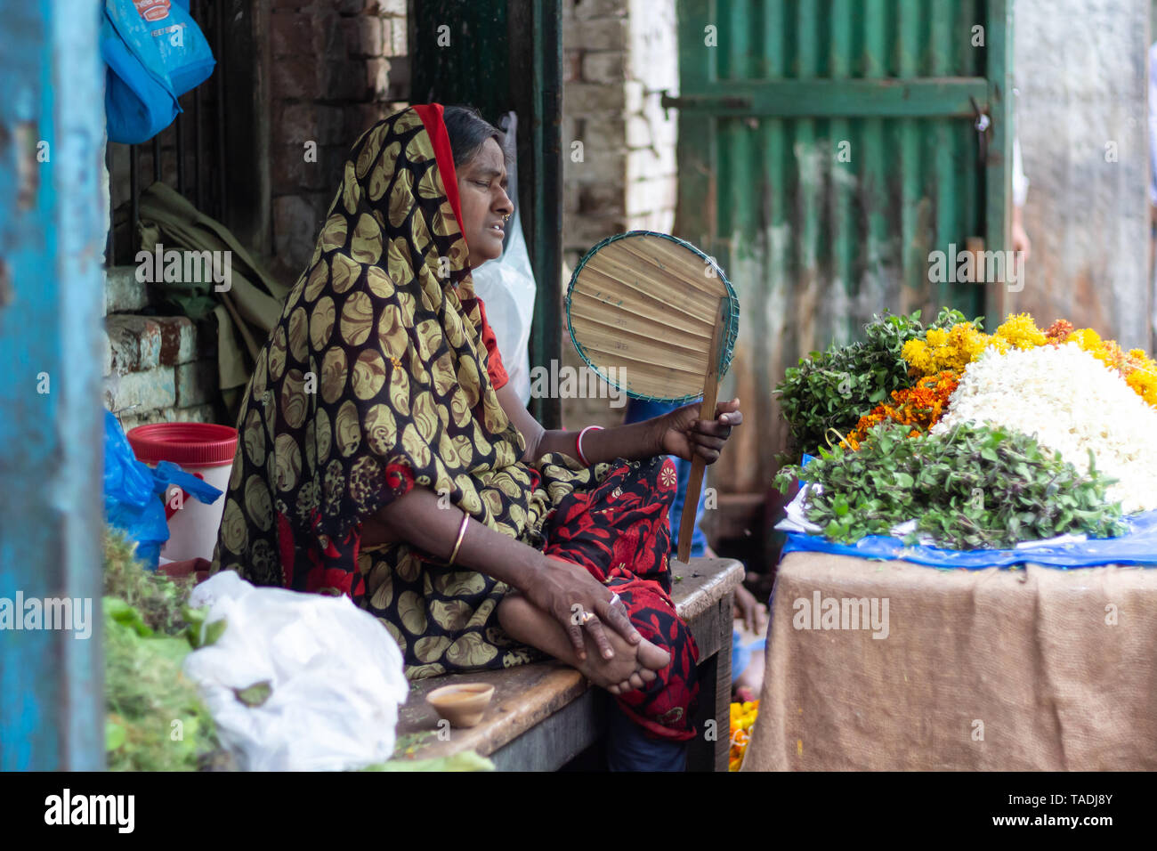 Il vecchio donna indiana esprimendo sgomento di alte temperature fanning se stessa nel mercato la vendita di piante e fiori. Foto Stock