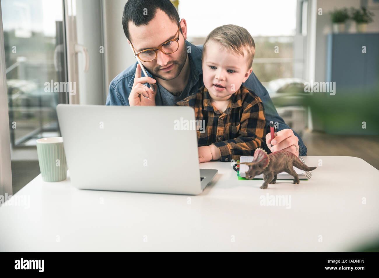 Padre lavora a tavola in casa ufficio con Figlio seduto sul suo giro Foto Stock