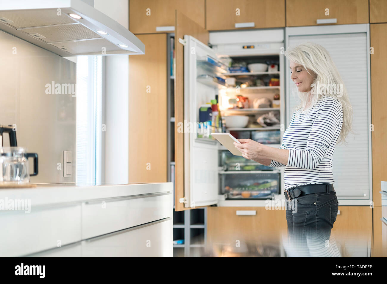 Contenuto donna matura in piedi in cucina a casa intelligente controllo frigo con tavoletta digitale Foto Stock