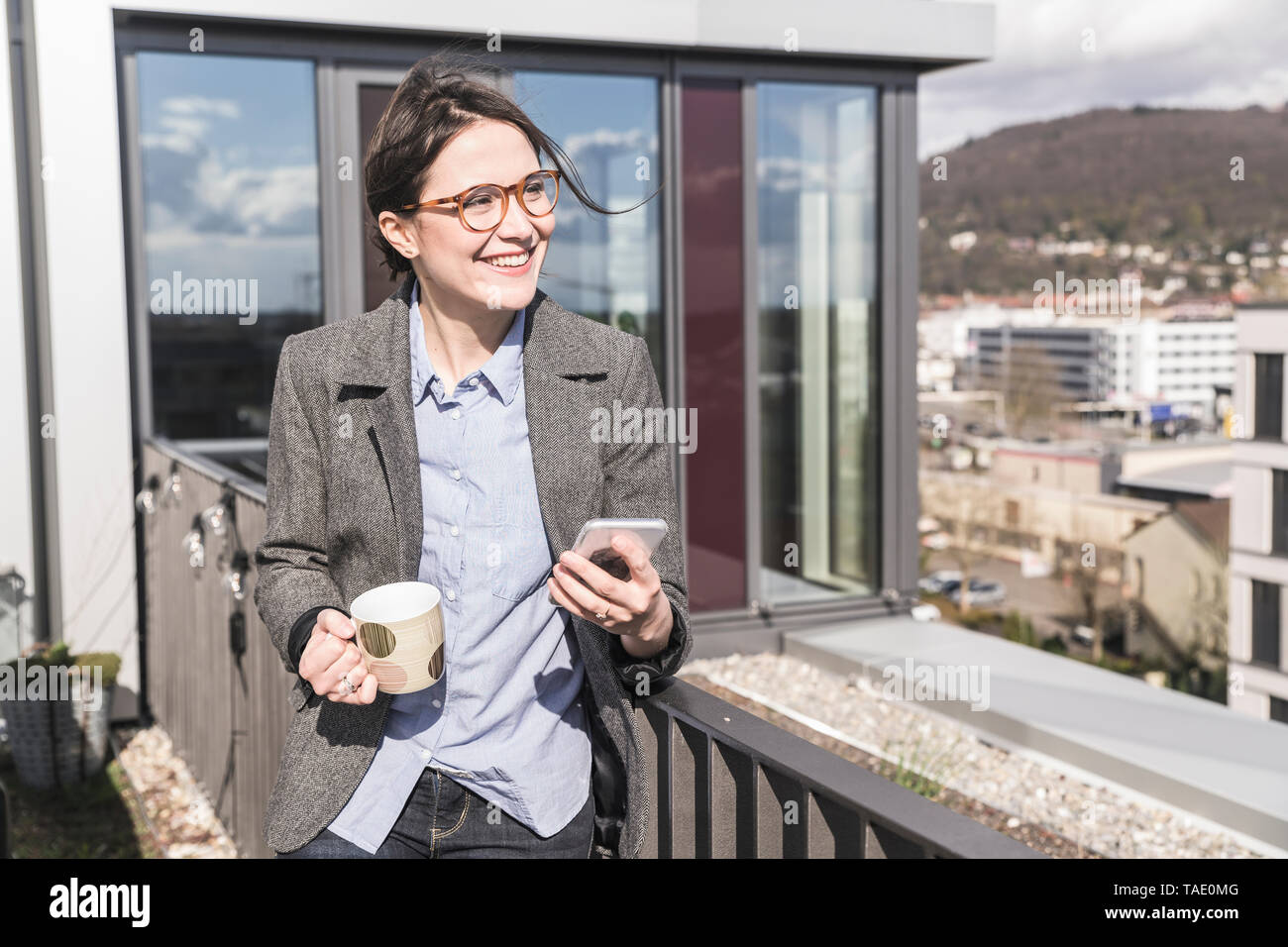 Imprenditrice sorridente con un telefono cellulare e la tazza di caffè sulla terrazza sul tetto Foto Stock